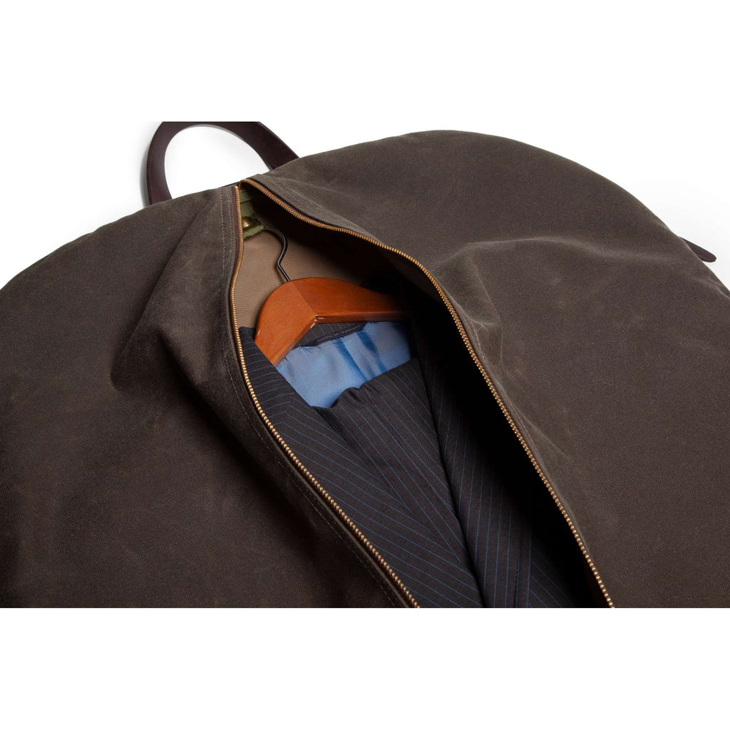 A dark brown waxed canvas garment bag with a zipper, shown open to reveal a blue pinstripe suit jacket and a wooden hanger inside, set against a plain white background.