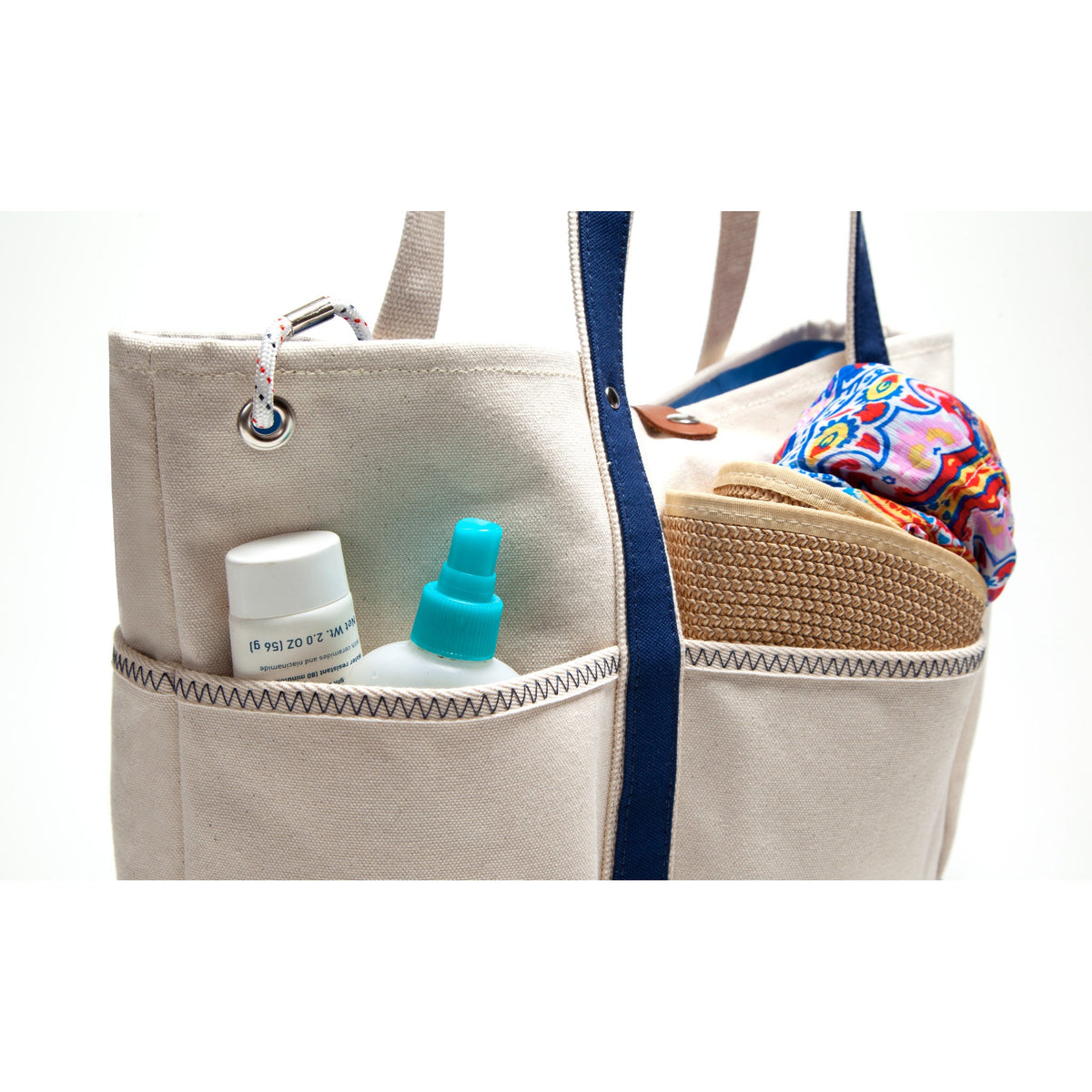 A beige canvas tote bag with navy blue straps and side pockets, shown holding travel items including a small white bottle with a turquoise sprayer, a white jar, a woven hat, and a colorful patterned cloth.