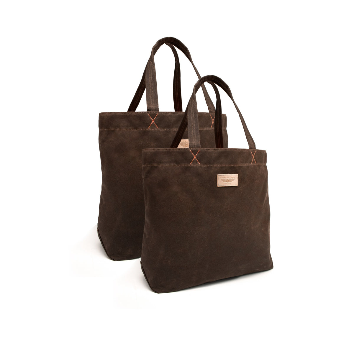 Two dark brown canvas tote bags with, featuring orange-brown contrast stitching and a small tan leather brand patch on the front, set against a plain white background.