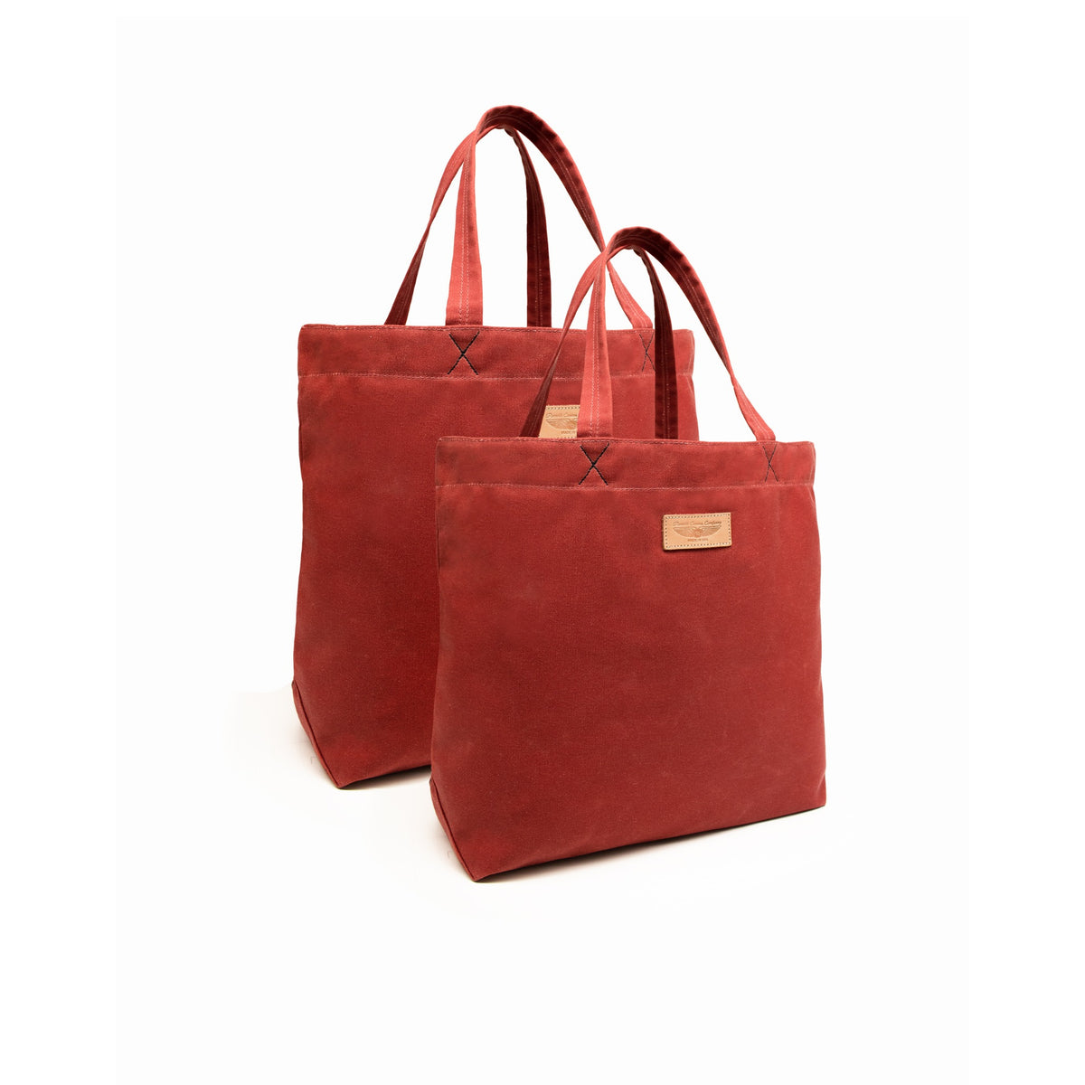 Two nantucket red canvas tote bags with matching handles, displayed on a clean white background. The front bag is slightly more prominent, showing a small brown leather brand patch on the front, while the second bag is positioned behind it.