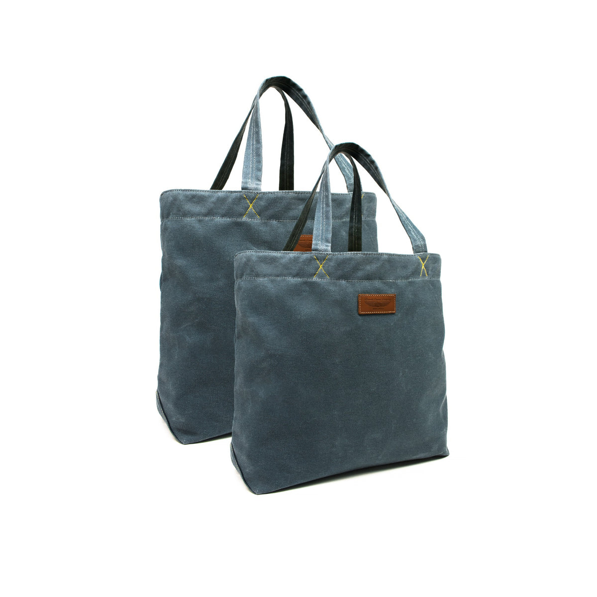 Two sanvas tote bags in a muted blue-gray color with black handles and a small brown leather brand patch on the front, shown on a plain white background.