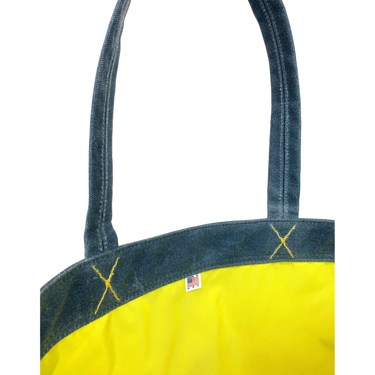 Close-up of a tote bag with dark blue denim-like handles and trim, a bright yellow body, yellow embroidered X details, and a small made in US flag tag near the top edge.