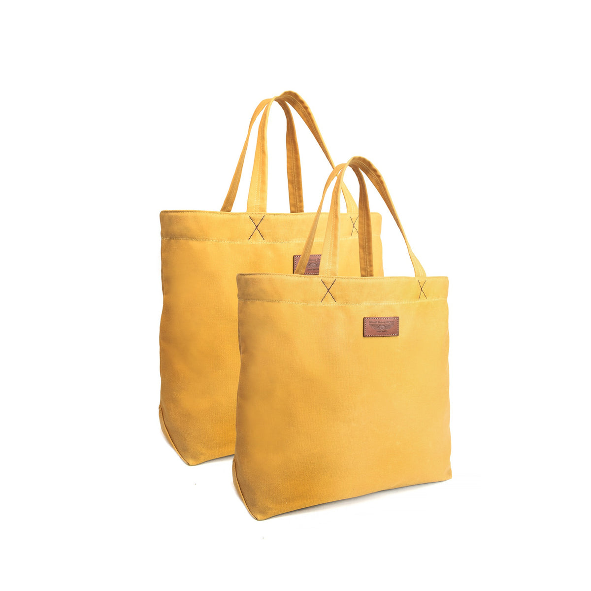 Two mustard yellow canvas tote bags with matching handles, displayed on a clean white background. The front bag is slightly more prominent, showing a small brown leather brand patch on the front, while the second bag is positioned behind it.
