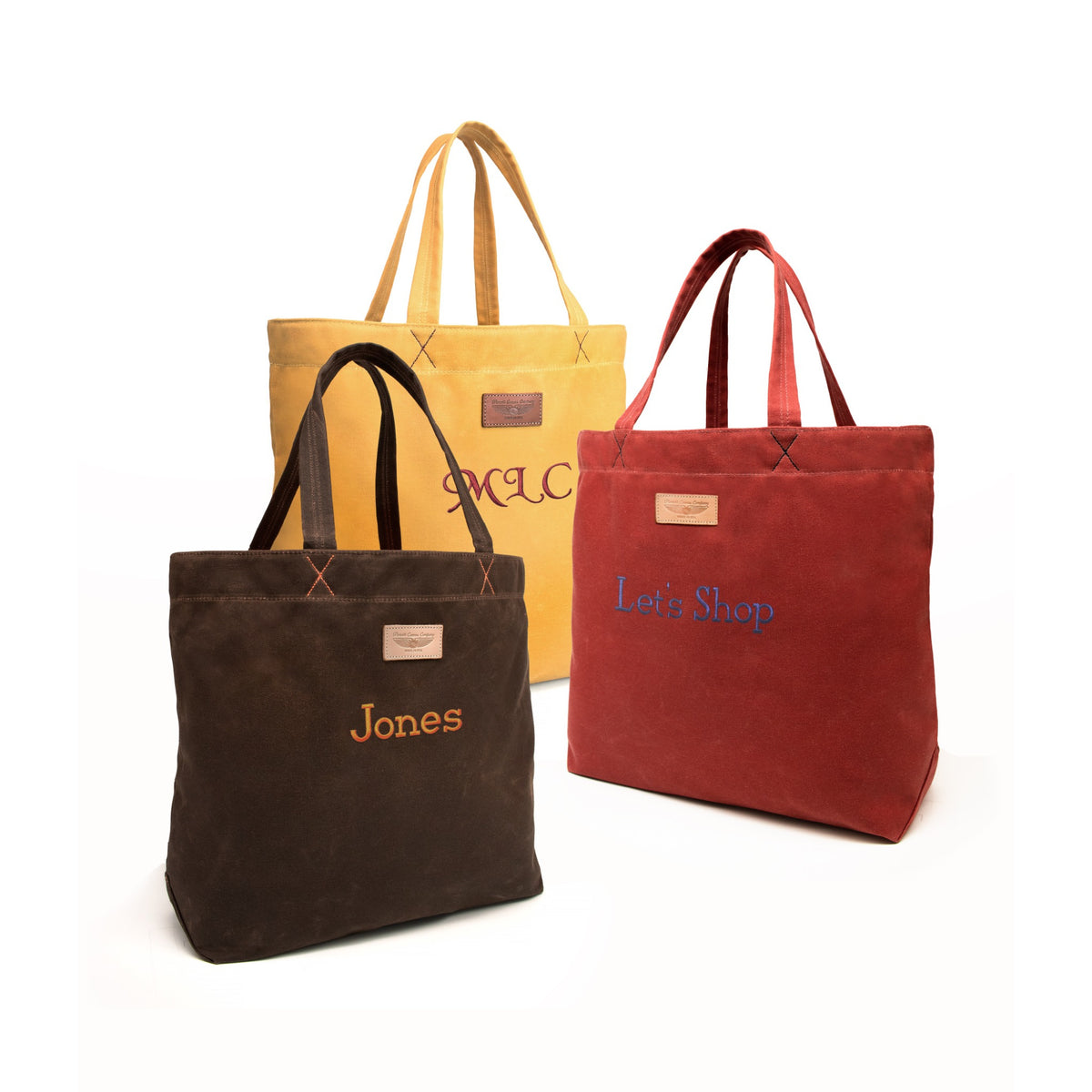 Three canvas tote bags in brown, mustard yellow, and rust red, each with embroidered text on the front and a small leather patch near the top, shown on a clean white background.
