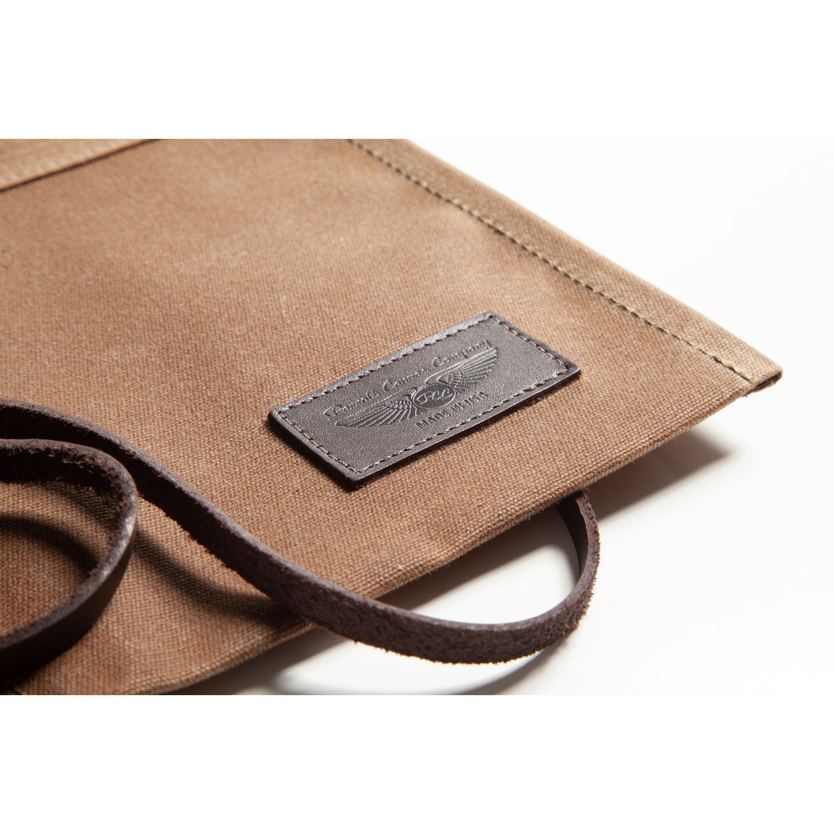 Close-up of a brown canvas bag with dark brown leather trim and a rectangular embossed leather brand patch on a white background.