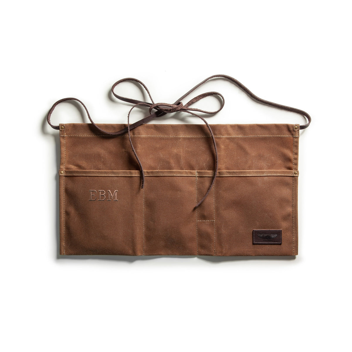 A brown canvas utility pouch with multiple pockets and a matching brown leather strap tied at the top, with a tan monogram of EBM and shown on a clean white background.