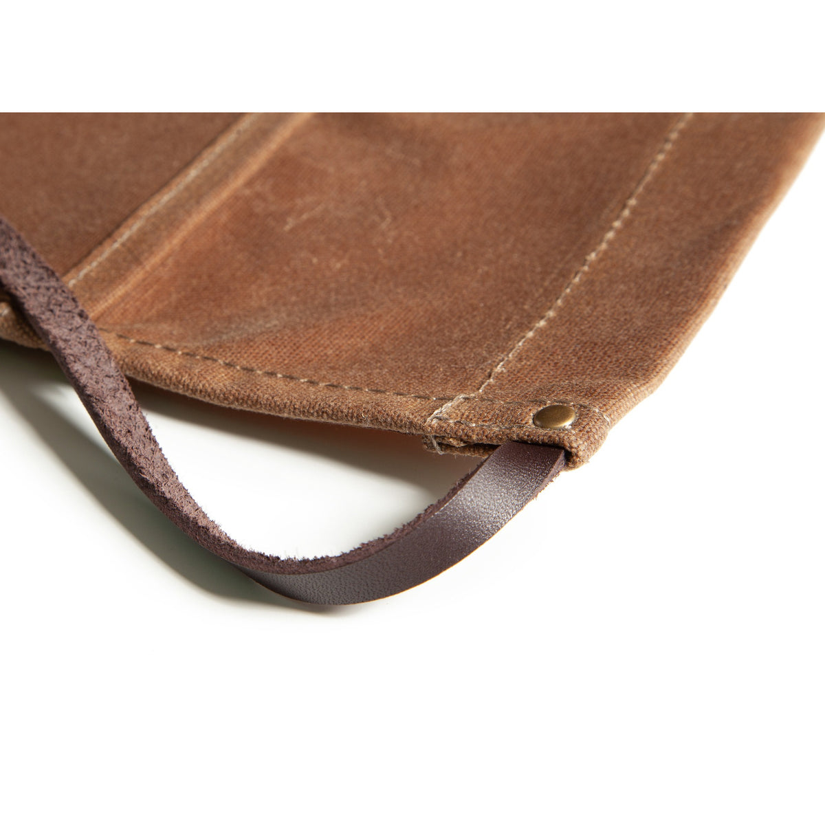 Close-up of a brown wax canvas apron corner with a leather strap and brass accent on a white background