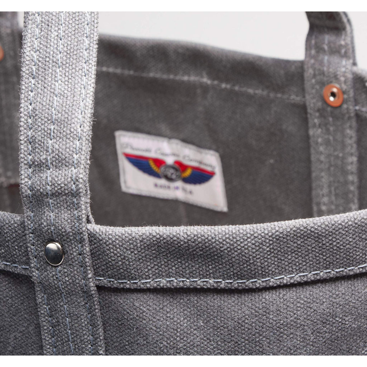 Close-up of a gray canvas tote bag showing the interior lining, metal rivets, and a sewn-on brand label that reads 'Parrott Canvas Company' with a red, blue and yellow parrott wings logo and 'Made in USA'.