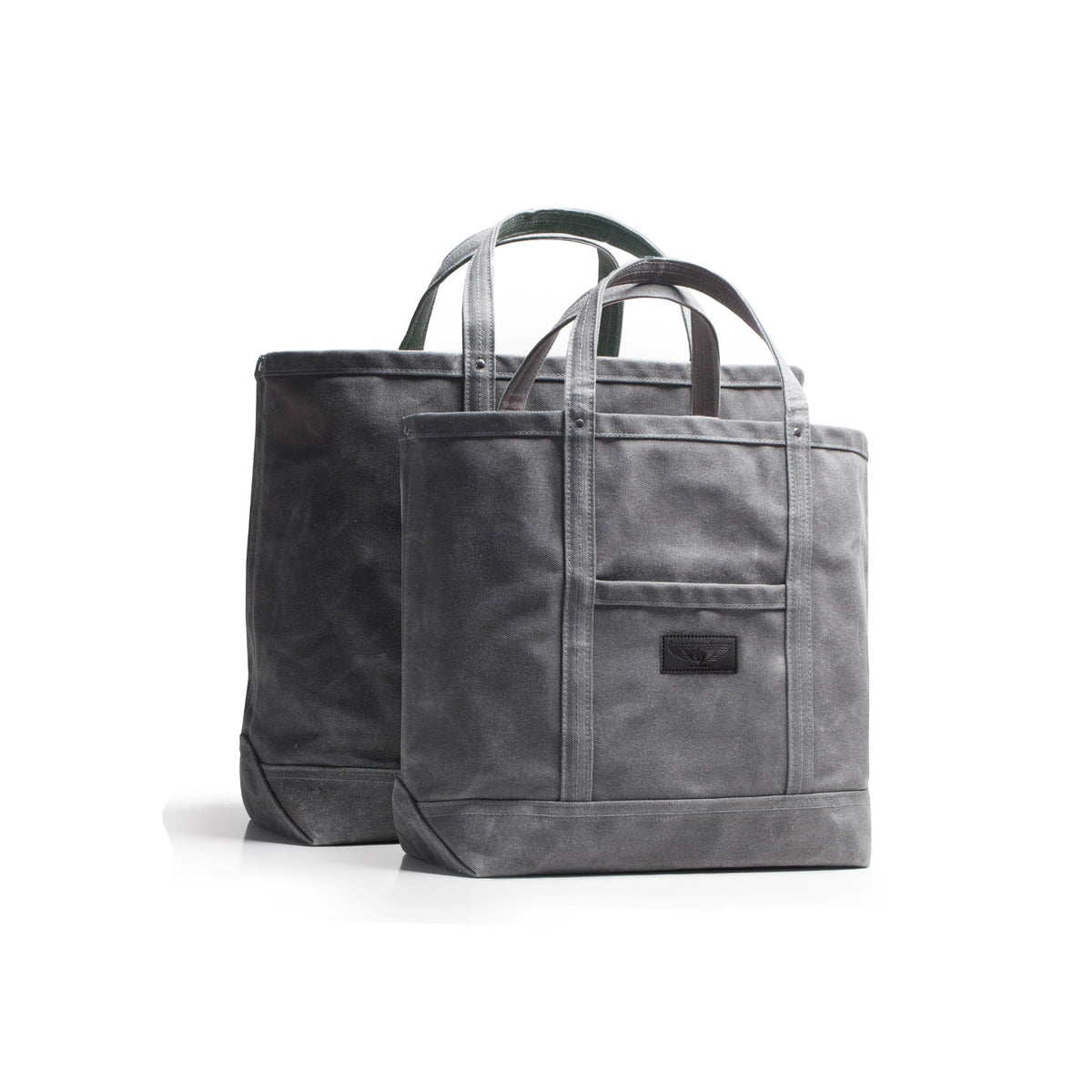 Two grey wax duck canvas tote bag and a small black leather brand patch on the front pocket, shown on a clean white background.