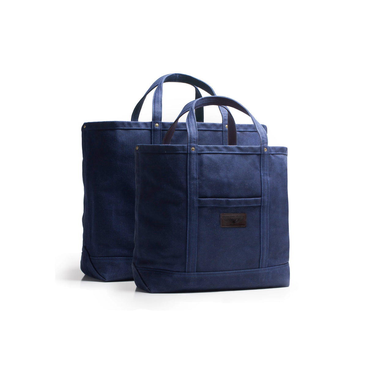 A navy blue waxed canvas tote bag with dual top handles and a black leather brand label and front pocket, shown in front of a second larger matching bag on a clean white background.